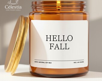 May include: A lit, amber-colored candle in a glass jar with a gold lid. The label reads "HELLO FALL." The candle is made with 100% natural soy wax and has a burn time of 40 hours. The jar is 9oz.