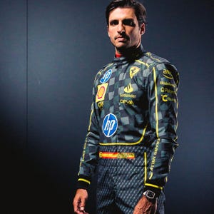 May include: A racing suit with a checkered pattern in shades of gray and green, accented with yellow stripes. Logos for HP, Shell, and Santander are visible. The suit features the name "CARLOS SAINZ" and the Spanish flag. A watch is worn on the wrist.