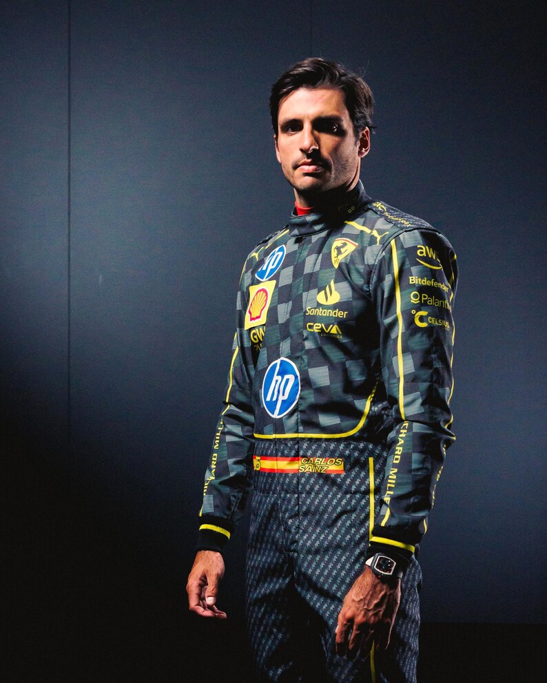 May include: A racing suit with a checkered pattern in shades of gray and green, accented with yellow stripes. Logos for HP, Shell, and Santander are visible. The suit features the name "CARLOS SAINZ" and the Spanish flag. A watch is worn on the wrist.