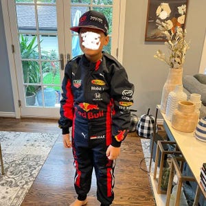 May include: A child wearing a black and red racing suit and a matching baseball cap. The suit features sponsor logos and the words "Red Bull" and "MAX". The cap has the words "Red Bull Racing" on it.