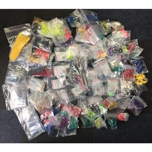 Puede incluir: Una colección de pequeños objetos coloridos en bolsas de plástico transparente. Los artículos incluyen accesorios amarillos, rojos, verdes y azules, junto con piezas de metal plateado. Las bolsas están dispuestas sobre una superficie oscura.