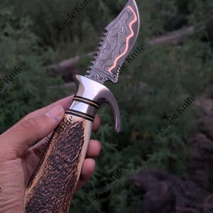 Könnte beinhalten: Ein feststehendes Messer mit einer gemusterten Damaststahlklinge, einer kupferfarbenen Wellenlinie, einem gezahnten Rücken, einem silberfarbenen Metallbacken mit gebogenem Parierelement und einem strukturierten braunen Hirschhorngriff.