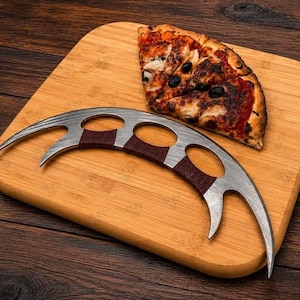 Könnte beinhalten: Ein halbmondförmiger Pizzaschneider aus Metall mit drei kreisförmigen Fingerlöchern und einem dunkelbraun umwickelten Griff. Die Klinge verfügt an beiden Enden über zwei scharfe, gebogene Spitzen, die zum Schneiden von Lebensmitteln dienen.