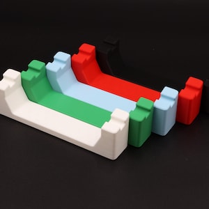 May include: A row of colourful phone stands in white, green, light blue, red, and black. Each stand has a curved design and a slot for holding a phone. The stands are arranged against a black background. Each stand is approximately 10 cm long.