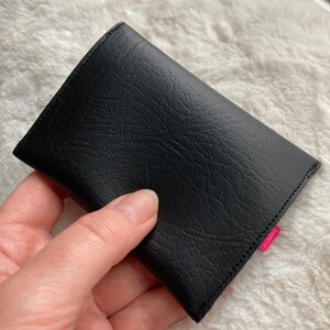 K&ouml;nnte beinhalten: Schwarze Ledergeldb&ouml;rse mit rosa Naht. Die Geldb&ouml;rse ist zusammengeklappt und wird in einer Hand gehalten.