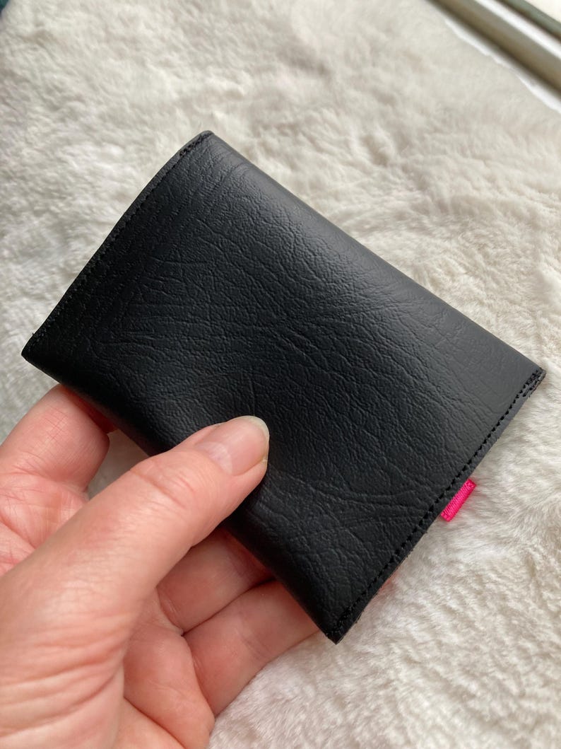 K&ouml;nnte beinhalten: Schwarze Ledergeldb&ouml;rse mit rosa Naht. Die Geldb&ouml;rse ist zusammengeklappt und wird in einer Hand gehalten.