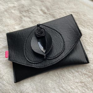 K&ouml;nnte beinhalten: Eine schwarze Leder-Clutch mit einer schwarzen Rosenverzierung.