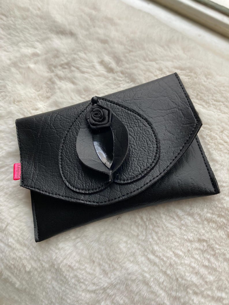 K&ouml;nnte beinhalten: Eine schwarze Leder-Clutch mit einer schwarzen Rosenverzierung.
