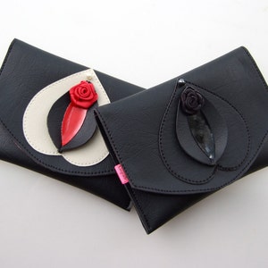 K&ouml;nnte beinhalten: Zwei schwarze Leder-Clutch-Taschen mit einem Blatt- und Blumendesign. Eine Clutch hat eine rote Blume und die andere eine schwarze Blume.