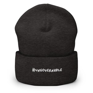 Könnte beinhalten: Dunkelgraue Beanie-Mütze mit dem weißen Text "#UNGOVERNABLE" auf dem umgeschlagenen Saum gestickt. Die Mütze ist aus weichem Strickmaterial gefertigt. Ein lässiges Accessoire für den Kopf.