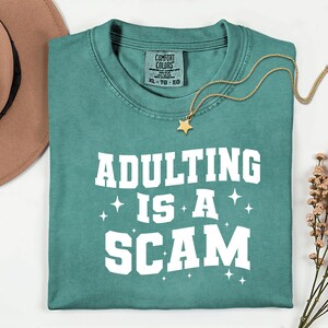 May include: Teal-colored t-shirt with the phrase "ADULTING IS A SCAM" in white block letters. A gold necklace with a star pendant rests on the shirt. The shirt is folded neatly, with a brown hat and flowers in the background.