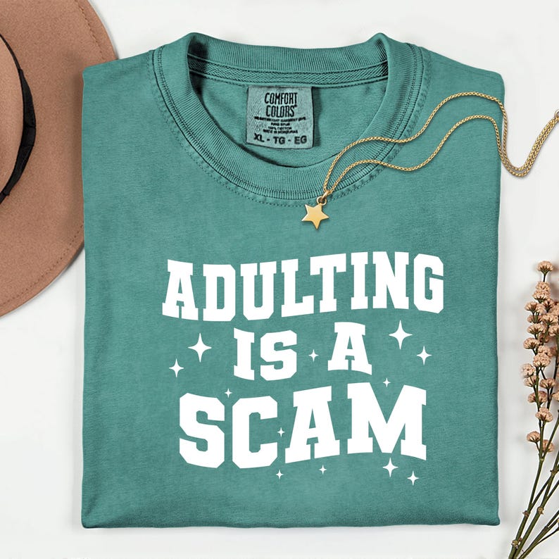 May include: Teal-colored t-shirt with the phrase "ADULTING IS A SCAM" in white block letters. A gold necklace with a star pendant rests on the shirt. The shirt is folded neatly, with a brown hat and flowers in the background.