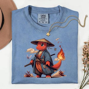 May include: A light blue XL Comfort Colors t-shirt featuring a cartoon character in a samurai outfit. The character is red with a flame on its tail, wearing a straw hat and holding two swords. A gold star necklace is draped across the shirt.