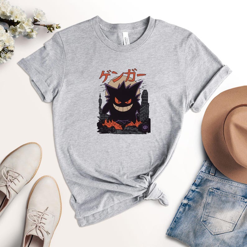 May include: A light gray t-shirt featuring a graphic of a purple monster with a sinister grin, set against a cityscape engulfed in flames. The Japanese text "ゲンガー" is above the monster. The shirt is made of a soft material.