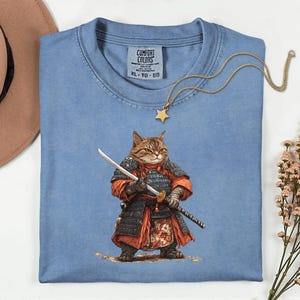 May include: A light blue Comfort Colors XL-TG-EG t-shirt featuring a detailed illustration of a cat dressed as a samurai, holding a katana and a wakizashi. A gold star pendant necklace rests on the shirt.