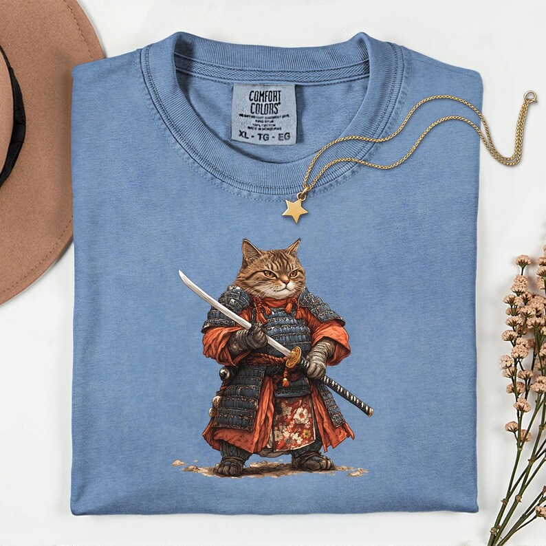 May include: A light blue Comfort Colors XL-TG-EG t-shirt featuring a detailed illustration of a cat dressed as a samurai, holding a katana and a wakizashi. A gold star pendant necklace rests on the shirt.