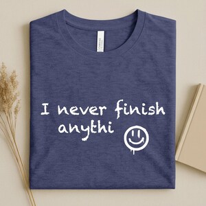 May include: A navy blue t-shirt with the white text "I never finish anythi" and a dripping smiley face. The shirt is folded and has a tag that says "Canvas". A book and dried plant are in the background.