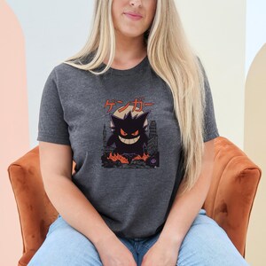 May include: Dark gray t-shirt featuring a black cartoon character with a city backdrop and flames. The Japanese text "ゲンガー" is above the character. The shirt is short-sleeved.