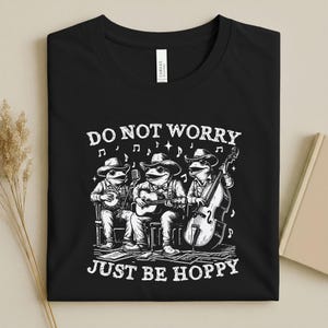 May include: Black t-shirt with a white graphic of three frog musicians playing instruments. The text "DO NOT WORRY JUST BE HOPPY" is printed above and below the frogs. The shirt is folded.