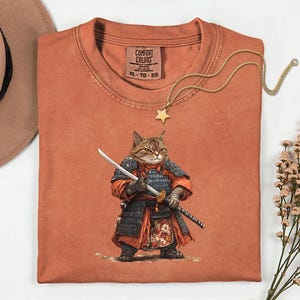 May include: A rust-colored Comfort Colors t-shirt featuring a detailed illustration of a cat dressed as a samurai, holding two swords. A gold star pendant necklace rests on the shirt. The shirt has a crew neck and short sleeves. The tag reads XL-TG-EG.