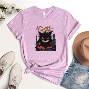 May include: A light purple t-shirt featuring a graphic of a black monster with a city backdrop and Japanese text. The monster has a wide grin and is surrounded by flames. The shirt is styled with a knot at the waist.
