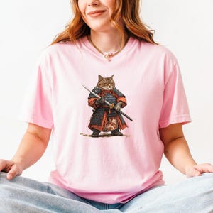 May include: Pink t-shirt featuring a detailed illustration of a cat dressed as a samurai warrior, complete with armor and a katana. The cat has brown and tan fur. The shirt is a solid pink color.
