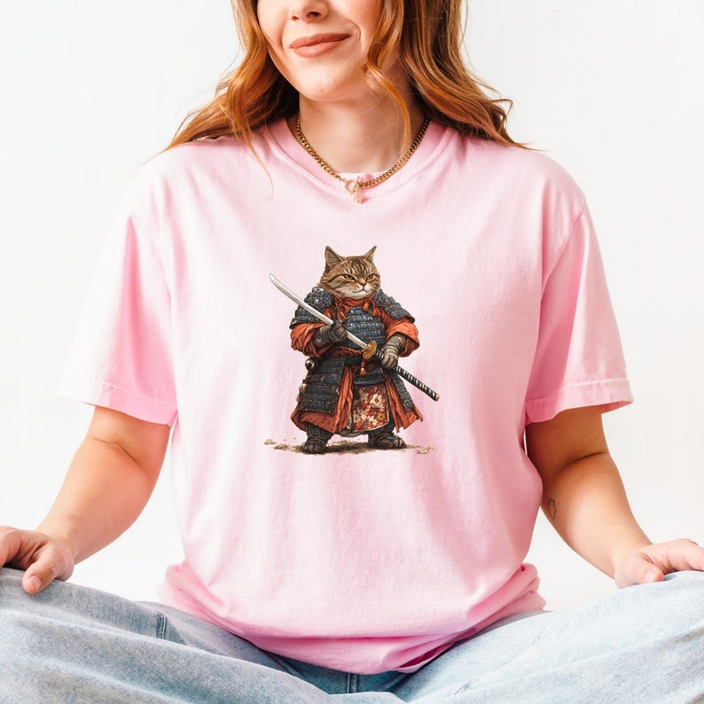 May include: Pink t-shirt featuring a detailed illustration of a cat dressed as a samurai warrior, complete with armor and a katana. The cat has brown and tan fur. The shirt is a solid pink color.