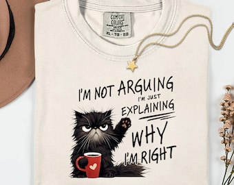 May include: Cream-colored t-shirt with the text "I'M NOT ARGUING I'M JUST EXPLAINING WHY I'M RIGHT" and a graphic of a grumpy black cat holding a red mug. A gold star pendant necklace is draped on the shirt.