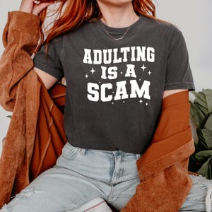May include: Dark gray t-shirt with the text "ADULTING IS A SCAM" in white block letters. The shirt has a crew neck and short sleeves. The person in the image is wearing a brown jacket and light blue jeans.