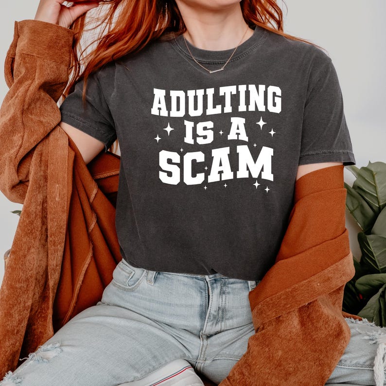 May include: Dark gray t-shirt with the text "ADULTING IS A SCAM" in white block letters. The shirt has a crew neck and short sleeves. The person in the image is wearing a brown jacket and light blue jeans.