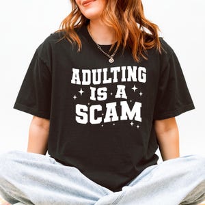 May include: Black t-shirt with the phrase "ADULTING IS A SCAM" in white block letters. The text is accented with small star graphics. The shirt is a casual style.