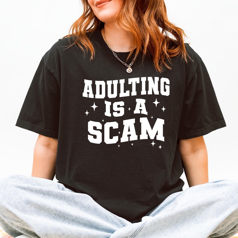 May include: Black t-shirt with the phrase "ADULTING IS A SCAM" in white block letters. The text is accented with small star graphics. The shirt is a casual style.