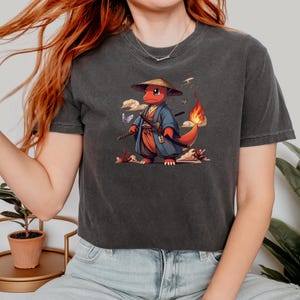 May include: Dark gray t-shirt featuring a red cartoon character in a samurai outfit, complete with a straw hat and swords. The character has a flame on its tail and is surrounded by clouds and butterflies. The design is printed on a soft, casual tee.