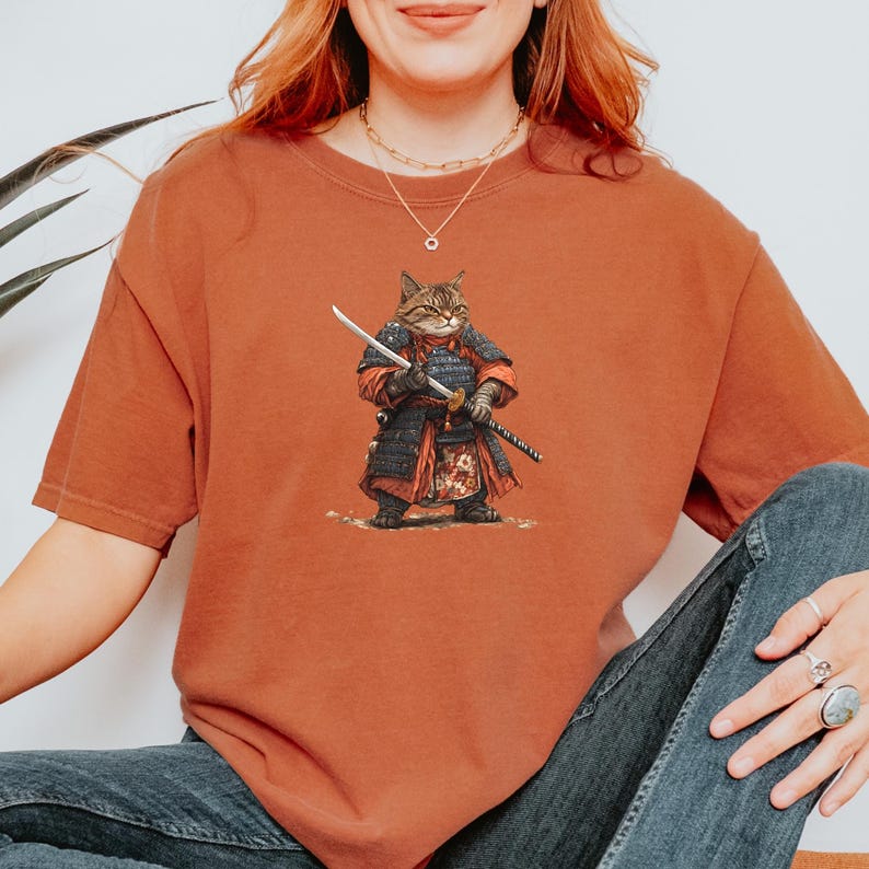 May include: Rust-colored t-shirt featuring a detailed illustration of a cat dressed as a samurai warrior, complete with armor and swords. The cat is depicted in a dynamic pose, holding a katana. The design is centered on the shirt.