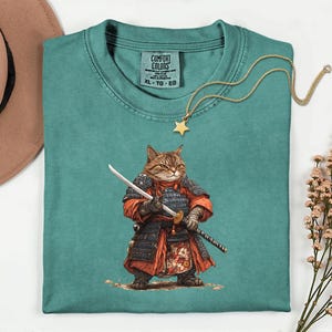 May include: Teal Comfort Colors t-shirt featuring a cat samurai illustration. The cat is depicted in traditional Japanese armor, holding a katana. A gold star pendant necklace rests on the shirt. The shirt has a tag that says "Comfort Colors XL-TG-EG".