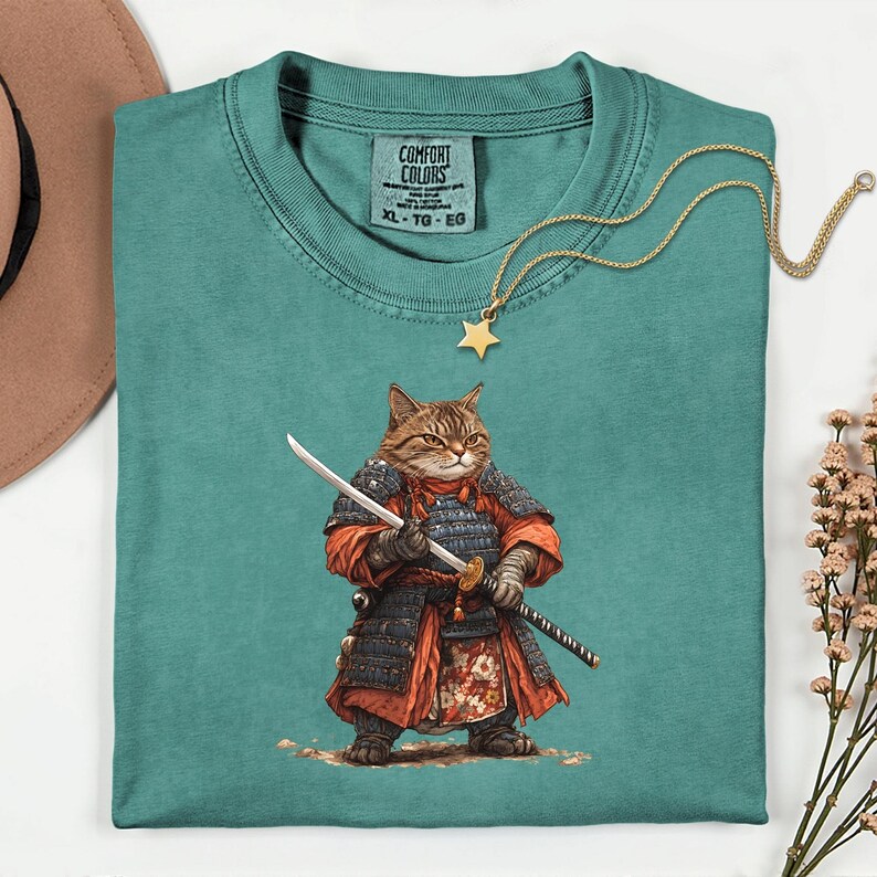 May include: Teal Comfort Colors t-shirt featuring a cat samurai illustration. The cat is depicted in traditional Japanese armor, holding a katana. A gold star pendant necklace rests on the shirt. The shirt has a tag that says "Comfort Colors XL-TG-EG".