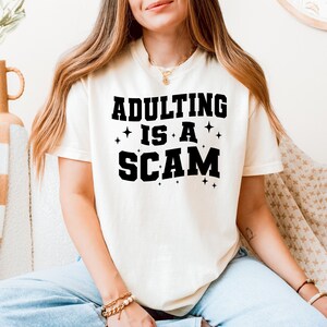 May include: Cream-colored t-shirt with the phrase "ADULTING IS A SCAM" in bold black letters. The text is accompanied by small star graphics. The shirt has short sleeves and a crew neck.