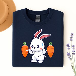 May include: Navy blue sweatshirt featuring a cartoon rabbit lifting two orange carrots as weights. The rabbit is white with pink-lined ears and a determined expression. The sweatshirt has a crew neck and a tag that reads "Gildan".