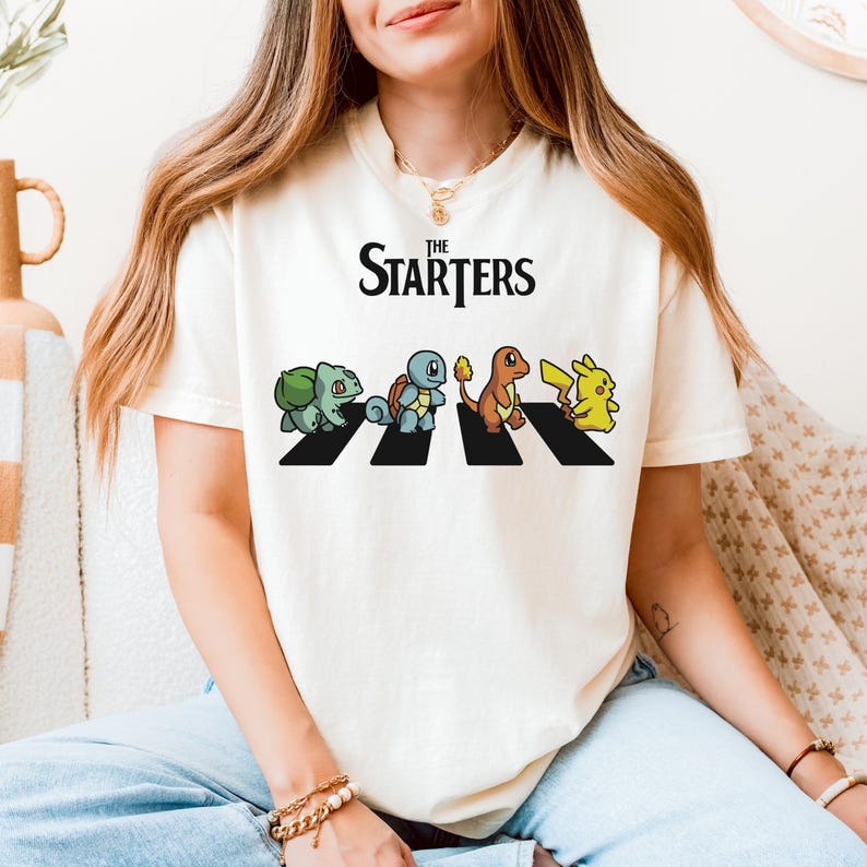 May include: Cream-coloured t-shirt with the text "The Starters" above a graphic of four cartoon characters crossing a black and white crosswalk. The characters are a green dinosaur, a blue turtle, an orange lizard, and a yellow mouse.