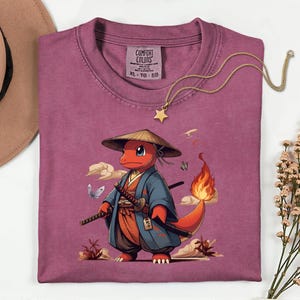 May include: A maroon Comfort Colors t-shirt featuring a cartoon character in a samurai outfit with two swords and a flame tail. The design includes clouds, butterflies, and a straw hat. A gold star pendant necklace is draped across the shirt.