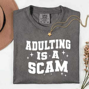 May include: A gray t-shirt with the phrase "ADULTING IS A SCAM" in white block letters. A gold star pendant necklace and a brown hat are also visible. The shirt has a Comfort Colors tag, size XL-TG-EG.
