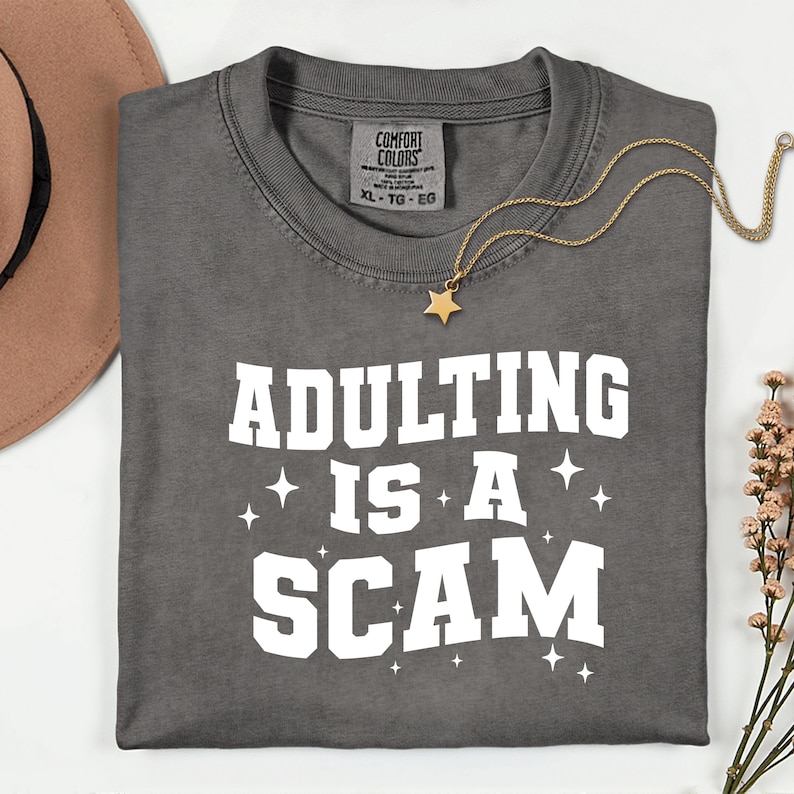 May include: A gray t-shirt with the phrase "ADULTING IS A SCAM" in white block letters. A gold star pendant necklace and a brown hat are also visible. The shirt has a Comfort Colors tag, size XL-TG-EG.