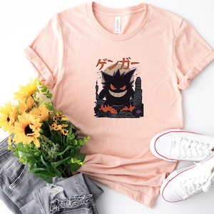 May include: A light pink t-shirt featuring a graphic of a black monster with the Japanese text "ゲンガー" above it. The monster is in front of a city skyline with flames. White sneakers and a bouquet of sunflowers are also in the image.
