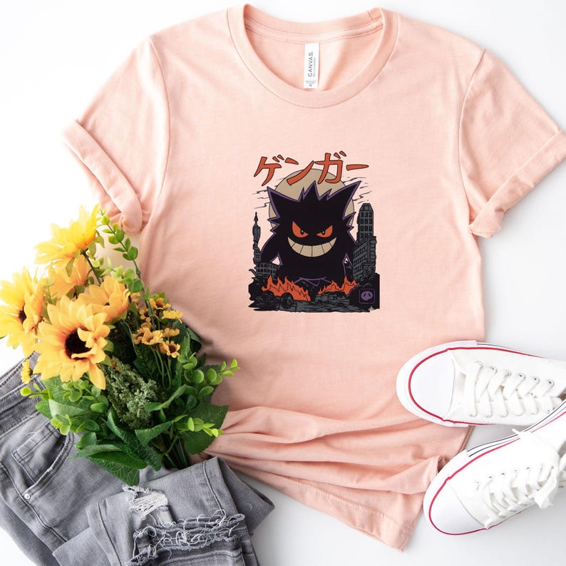 May include: A light pink t-shirt featuring a graphic of a black monster with the Japanese text "ゲンガー" above it. The monster is in front of a city skyline with flames. White sneakers and a bouquet of sunflowers are also in the image.