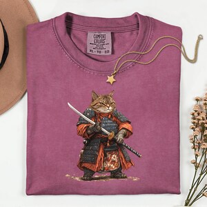 May include: A maroon Comfort Colors t-shirt featuring a detailed illustration of a cat dressed as a samurai, holding a katana and a wakizashi. The shirt has a crew neck and the size is XL-TG-EG. A gold star pendant necklace is draped across the shirt.