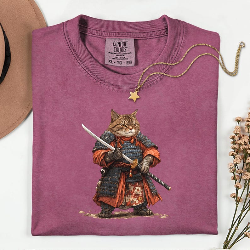 May include: A maroon Comfort Colors t-shirt featuring a detailed illustration of a cat dressed as a samurai, holding a katana and a wakizashi. The shirt has a crew neck and the size is XL-TG-EG. A gold star pendant necklace is draped across the shirt.