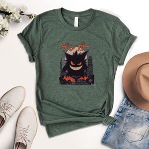 May include: Green t-shirt featuring a black cartoon character with a city backdrop and Japanese text. The character has a large grin and is surrounded by flames. A brown hat, white shoes, and blue jeans are visible.