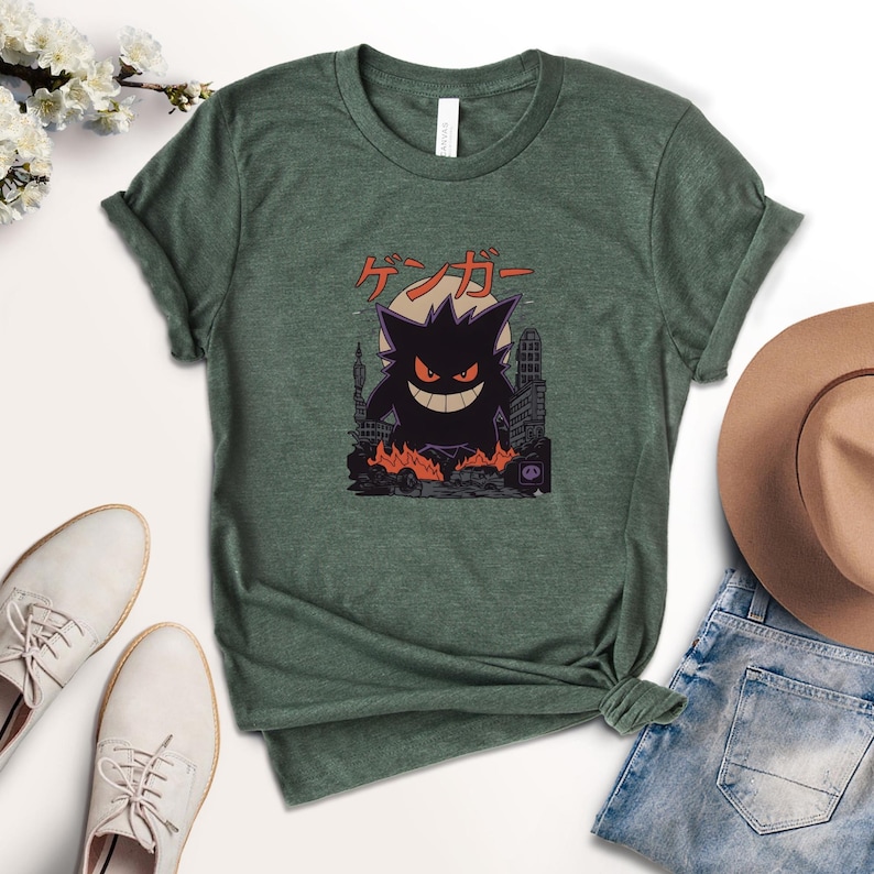 May include: Green t-shirt featuring a black cartoon character with a city backdrop and Japanese text. The character has a large grin and is surrounded by flames. A brown hat, white shoes, and blue jeans are visible.