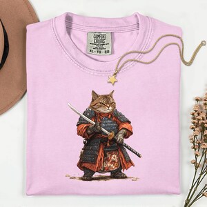 May include: A light pink Comfort Colors XL t-shirt featuring a detailed illustration of a cat dressed as a samurai, holding a katana. The cat is wearing armor and a red and black outfit. A gold star pendant necklace is draped across the shirt.