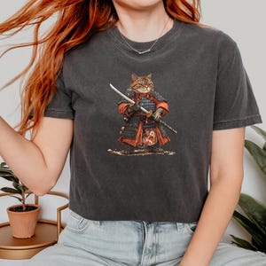 May include: Dark gray t-shirt featuring a cartoon illustration of a cat dressed as a samurai, holding a katana. The cat is wearing red and black armor. The design is centered on the shirt.